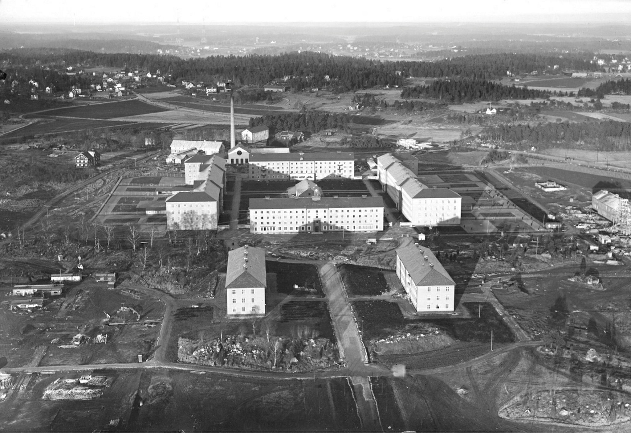 Svartvitt foto på flera stora byggnader sedda från luften. Byggnaderna liknar varandra, är avlånga och har många fönster på rad och formar tillsammans en kvadrat. Runt om byggnaderna finns åkrar, träd och andra mindre hus.