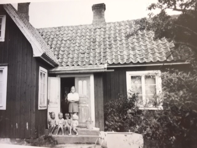 Svartvitt foto med en stuga. Dörren till stugan är öppen. Det är sommar. På trappen sitter fyra barn som är ganska nära varandra i ålder. Bakom dem står en kvinna.