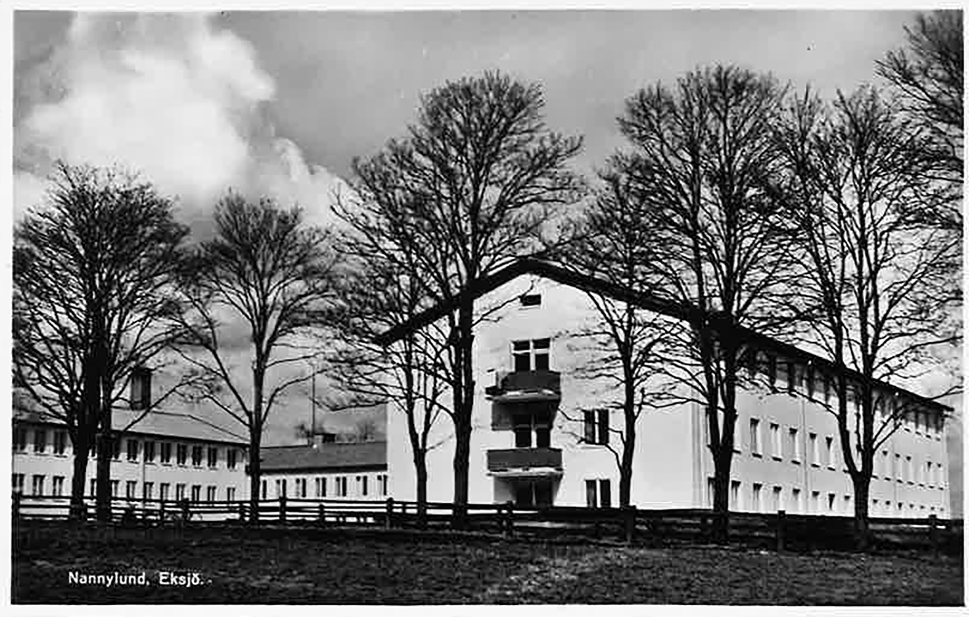 Svartvitt foto med tre byggnader. Utsidorna har många likadana fönster i rader. Husen är två och tre våningar höga och avlånga. Det finns ett staket runt byggnaderna. Längst fram på bilden finns några träd.